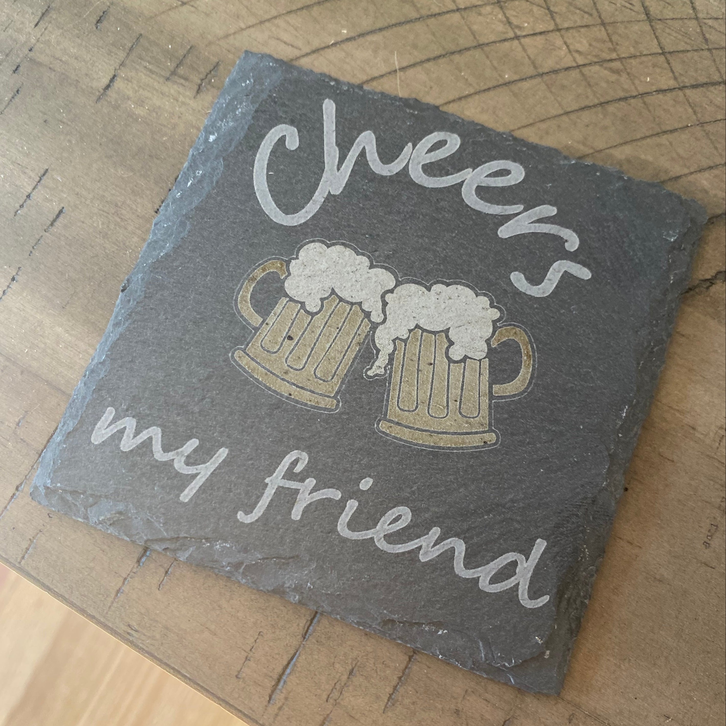 Slate coaster with 'Cheers my friends' text and beer mug design on a wooden surface